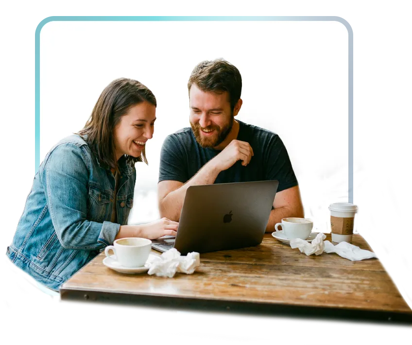 A woman in a denim jacket and a bearded man in a gray t-shirt sit together at a rustic wooden table, laughing as they look at a MacBook laptop. Coffee cups and crumpled napkins are on the table, suggesting a casual, collaborative work session in a cafe setting.