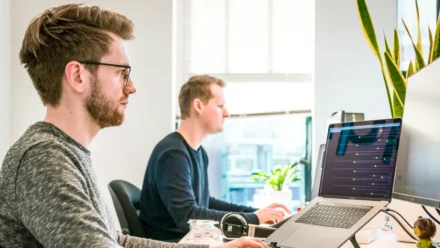 Two IT professionals collaborating at desks with MacBooks, reviewing Apple device management dashboard for MSPs scaling secure fleets without extra headcount
