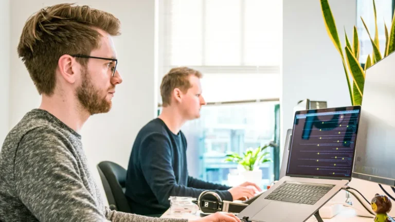 Two IT professionals collaborating at desks with MacBooks, reviewing Apple device management dashboard for MSPs scaling secure fleets without extra headcount