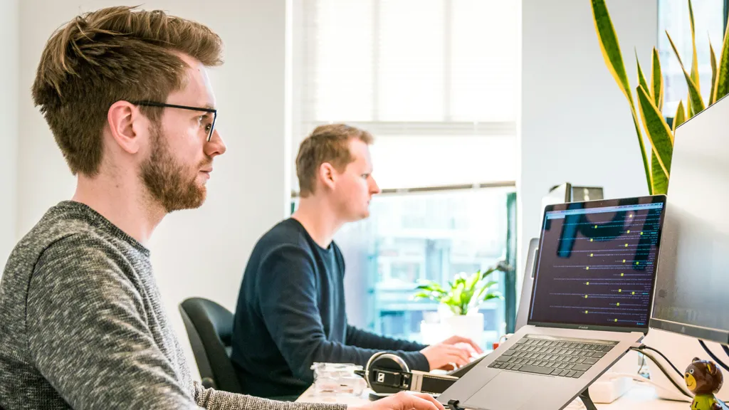 Two IT professionals collaborating at desks with MacBooks, reviewing Apple device management dashboard for MSPs scaling secure fleets without extra headcount