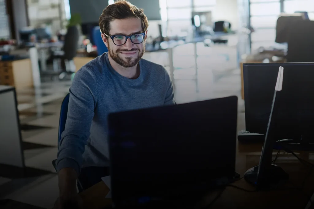 A smiling man with glasses and a beard works at a laptop in a modern open-plan office, with a second monitor visible beside him.
