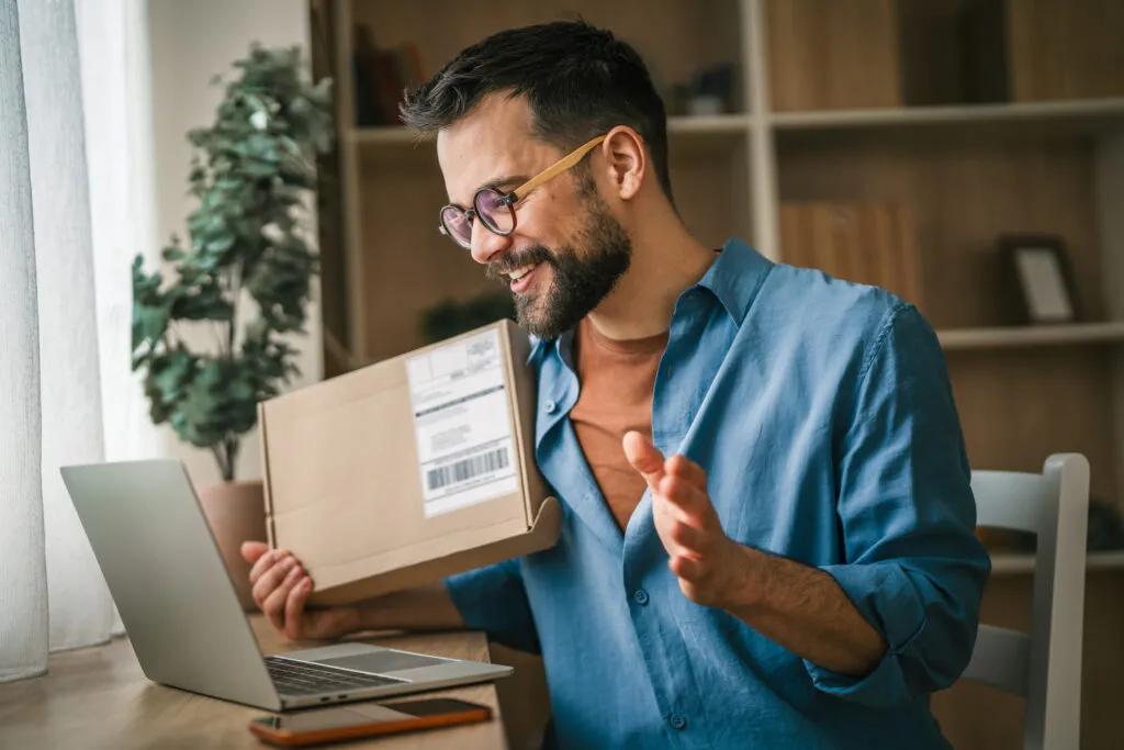 A smiling man with glasses and a beard holds a cardboard shipping package while sitting at a desk with a laptop open in front of him, appearing pleased with a recent delivery.