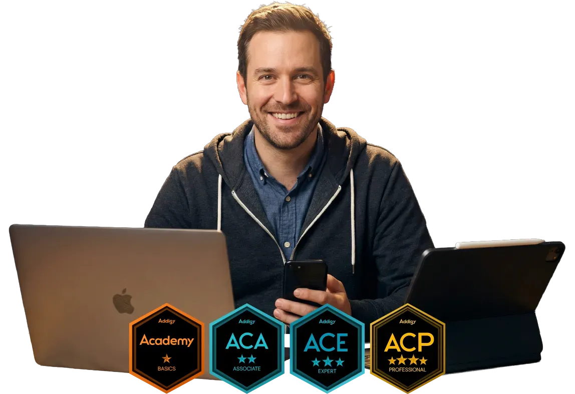 A smiling man in a gray hoodie over a blue denim shirt sits at a desk holding a smartphone, flanked by an Apple MacBook laptop on the left and an iPad with a keyboard case on the right. In the foreground are four Addigy certification badges: Academy (one star), ACA – Associate (two stars), ACE – Expert (three stars), and ACP – Professional (four stars).
