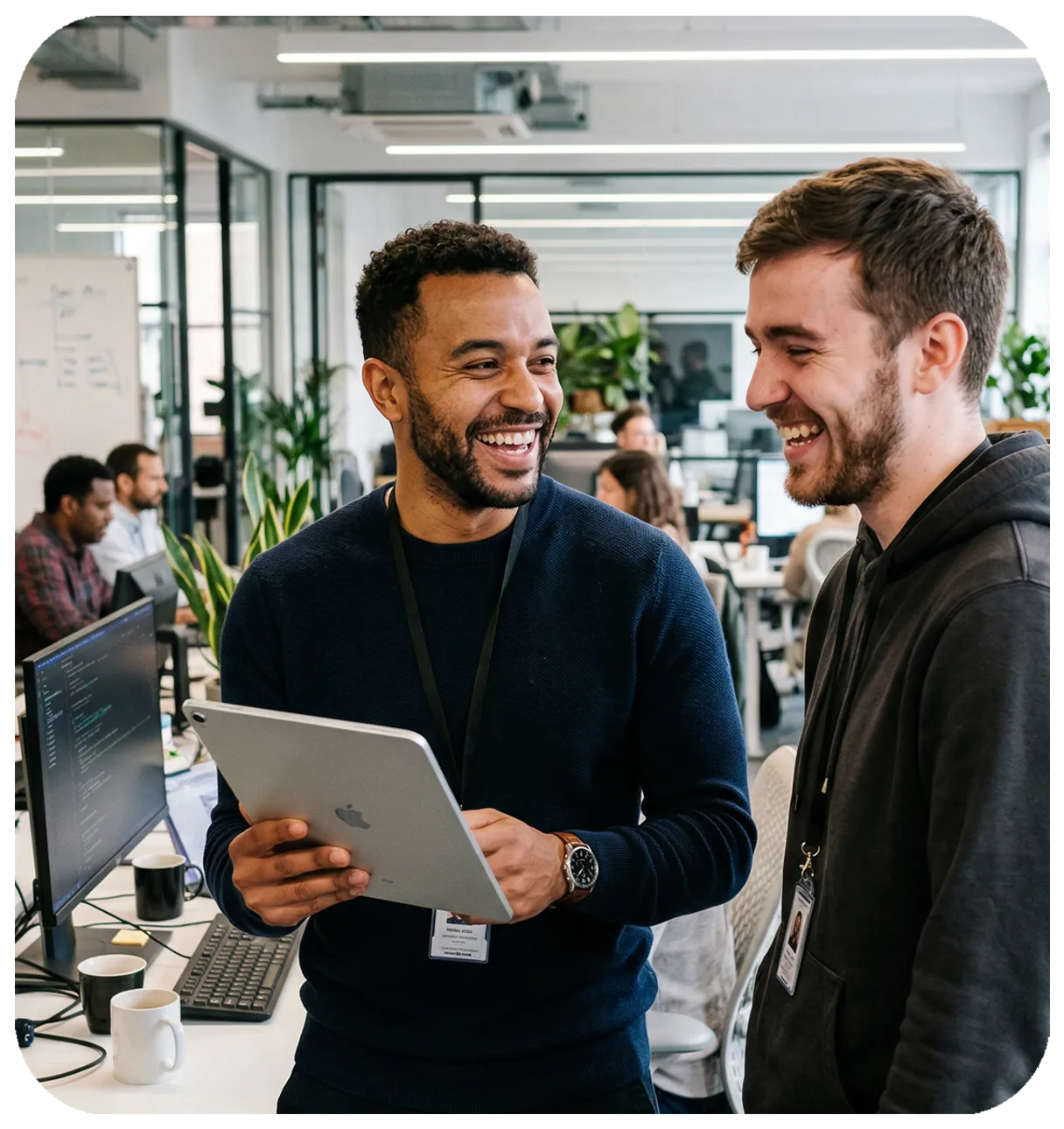 Two IT professionals laughing together in a modern office, one holding an iPad and showing the screen to a colleague